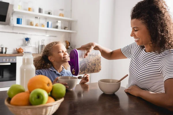 Selektiver Fokus der jungen afrikanisch-amerikanischen Frau im gestreiften T-Shirt, die Cornflakes in Schüssel neben aufgeregter Tochter gießt — Stockfoto