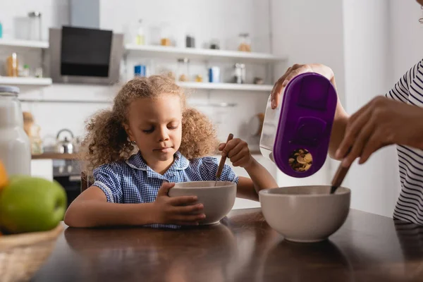 Vista parcial de la mamá afroamericana vertiendo copos de maíz en un tazón cerca de su hija desayunando - foto de stock