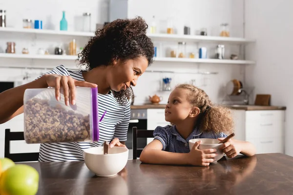 Young african american woman looking at daughter while pouring cornflakes into bowl for breakfast — Stock Photo