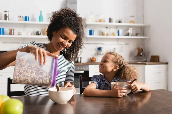 Jovem mulher afro-americana derramando flocos de milho para o café da manhã em tigela perto da filha — Fotografia de Stock