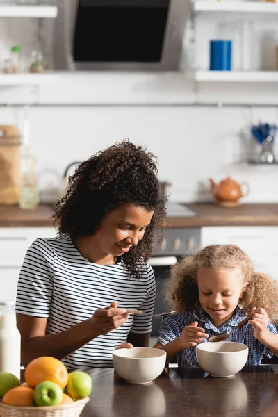 Madre afroamericana y su hija sosteniendo cucharas cerca de cuencos con desayuno - foto de stock