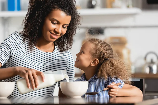 Giovane donna afroamericana in t-shirt a righe versando latte fresco in ciotola vicino figlia tenendo cucchiaio — Foto stock