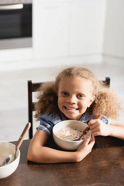 Vista de ángulo alto de la chica afroamericana emocionada mirando a la cámara mientras sostiene la cuchara cerca del tazón con leche y copos de maíz - foto de stock