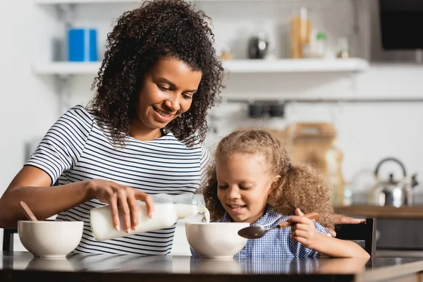 Молодая африканская американская мать в полосатой футболке наливает молоко в миску рядом с дочерью, держа ложку — стоковое фото