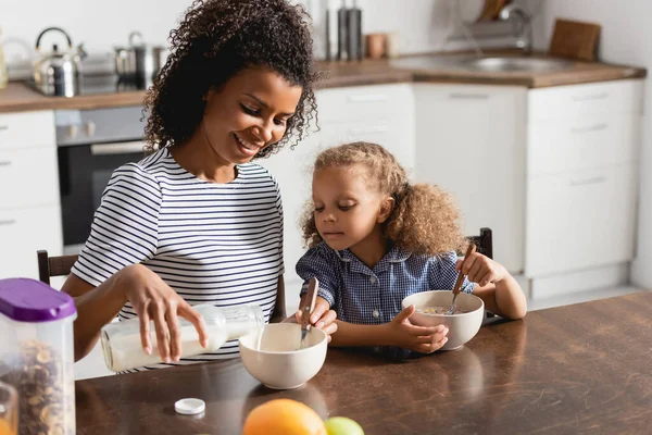 Afrikanisch-amerikanische Mutter in gestreiftem T-Shirt gießt Milch in Schüssel, während sie mit Tochter frühstückt — Stockfoto