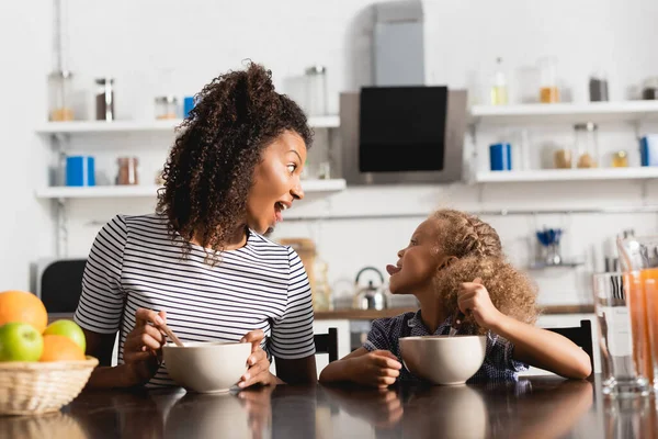 Excitada mujer afroamericana en camiseta a rayas mirando a su hija sobresaliendo de la lengua durante el desayuno en la cocina - foto de stock