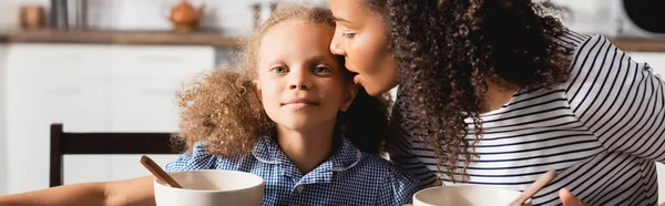 Colpo panoramico di donna in t-shirt a righe sussurrando in orecchio di figlia durante la colazione — Foto stock