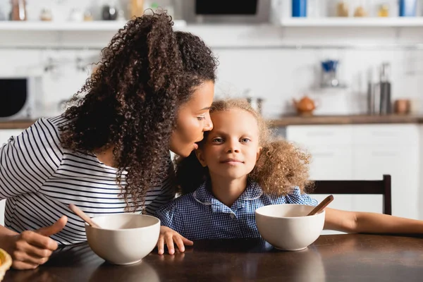 Mujer afroamericana en camiseta a rayas susurrando al oído de su hija durante el desayuno - foto de stock