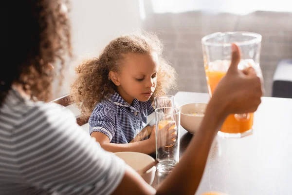 Селективное внимание матери, держащей кувшин со свежим апельсиновым соком рядом с африканской американской дочерью — стоковое фото