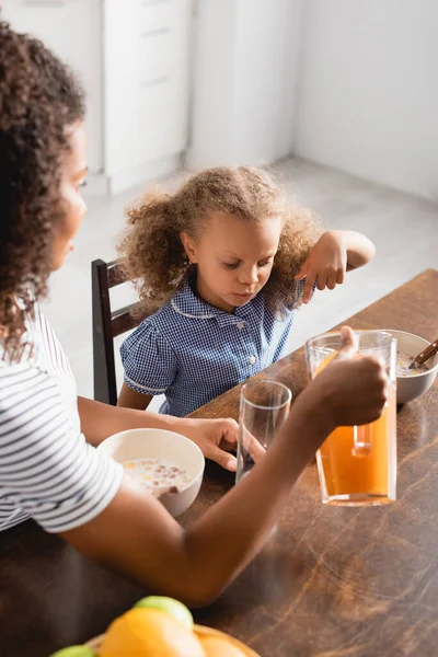 Selective focus of mother holding jug with orange juice near african american daughter pointing with finger during breakfast — Stock Photo