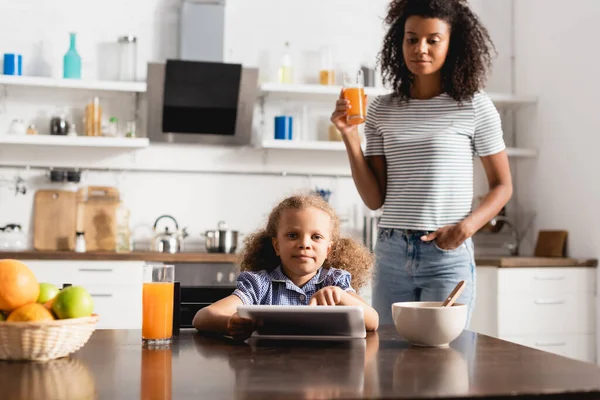 Jeune femme afro-américaine tenant du jus d'orange tout en se tenant la main dans la poche près de la fille à l'aide d'une tablette numérique — Photo de stock