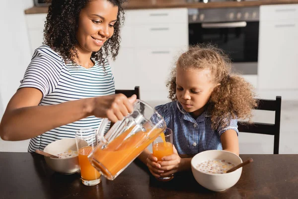 Joven afroamericana americana mujer en rayas camiseta verter jugo de naranja fresca en vasos cerca de hija durante el desayuno - foto de stock