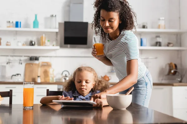 Madre afroamericana sosteniendo jugo de naranja y tocando la tableta digital cerca del niño concentrado en la cocina - foto de stock
