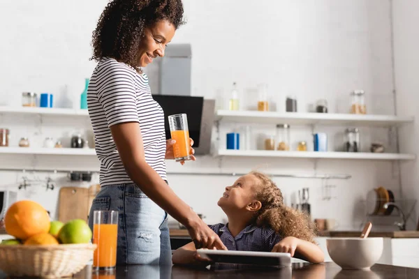 Joven afroamericana mujer sosteniendo jugo de naranja y tocando tableta digital cerca de hija en cocina - foto de stock