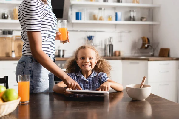 Teilbild einer afrikanisch-amerikanischen Frau im gestreiften T-Shirt mit Orangensaft und anrührendem digitalen Tablet in der Nähe der aufgeregten Tochter — Stockfoto