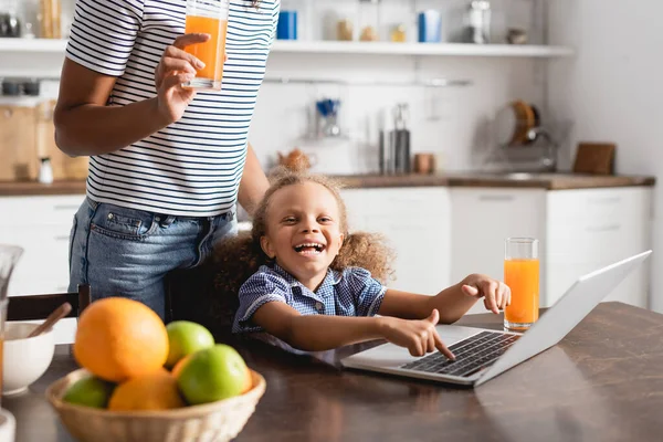 Vista ritagliata di donna con vetro di succo d'arancia vicino eccitata figlia afro-americana utilizzando il computer portatile in cucina — Foto stock