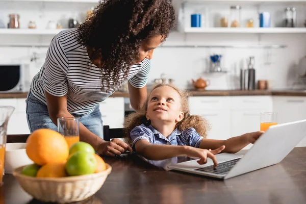 Enfoque selectivo de la madre y la hija afroamericana mirándose cerca de la computadora portátil en la cocina - foto de stock