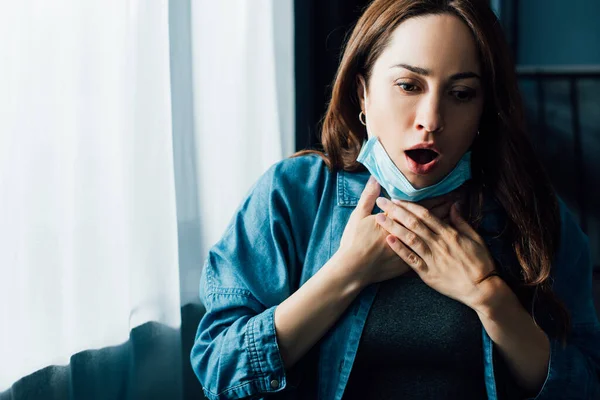 Mujer asustada con la boca abierta en máscara médica tocando la garganta - foto de stock