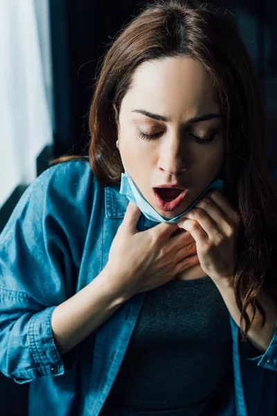 Femme effrayée avec les yeux fermés et la bouche ouverte dans le masque médical touchant la gorge — Photo de stock