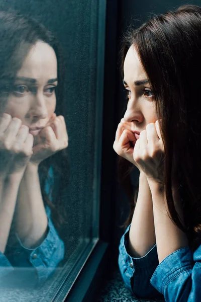 Triste morena mujer tocando la cara mientras mira a la ventana - foto de stock