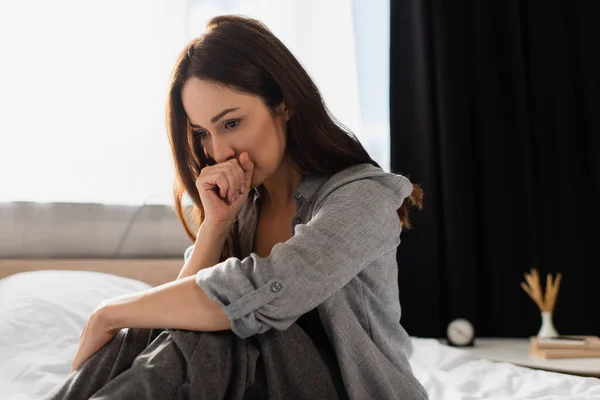 Depressive brünette Frau, die den Mund bedeckt, während sie zu Hause auf dem Bett sitzt — Stockfoto