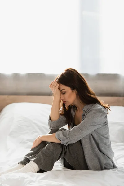 Mujer morena deprimida con los ojos cerrados sentado en la cama y tocando la cara en casa - foto de stock