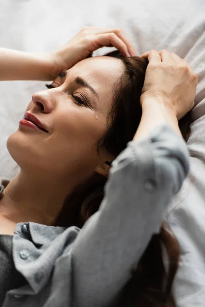 Розчарована і брюнетка жінка з закритими очима торкається волосся під час плачу на ліжку — стокове фото