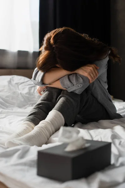 Messa a fuoco selettiva di depresso bruna donna che copre il viso mentre seduto sul letto vicino scatola di tessuto — Foto stock