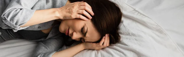 Panoramic shot of brunette woman having migraine, touching head and lying on bed — Stock Photo