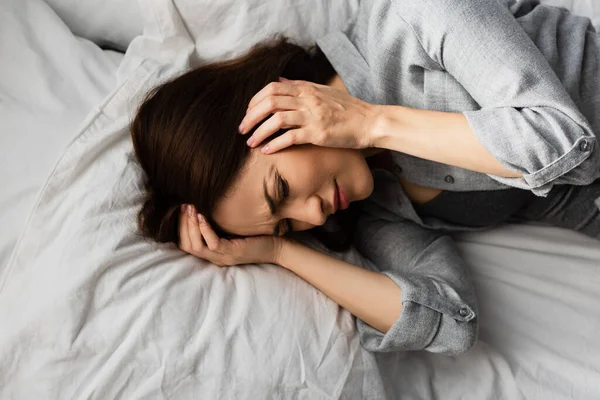 Top view of tired woman having migraine, touching head and lying on bed — Stock Photo