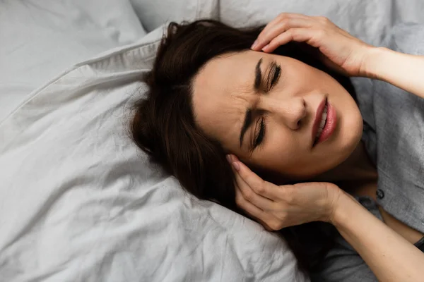 Top view of brunette woman having migraine, touching head and lying on bed — Stock Photo