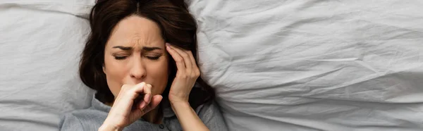 Horizontal crop brunette woman suffering from pain and touching head while lying on bed — Stock Photo