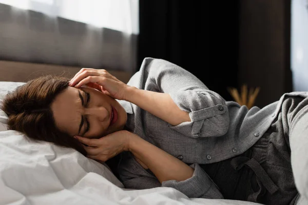 Втомлена брюнетка жінка страждає від болю, торкаючись голови і лежачи на ліжку — стокове фото