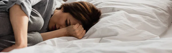 Concepto panorámico de mujer morena que sufre de dolor y tocando la cabeza mientras está acostada en la cama - foto de stock