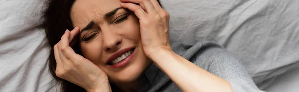 Panoramic shot of brunette woman suffering from pain and touching head while lying on bed — Stock Photo