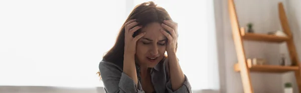 Raccolto panoramico di donna bruna con gli occhi chiusi che soffrono di dolore e toccano la testa a casa — Stock Photo