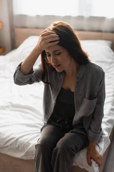 Tired woman with closed eyes suffering from pain and touching head while sitting on bed at home — Stock Photo