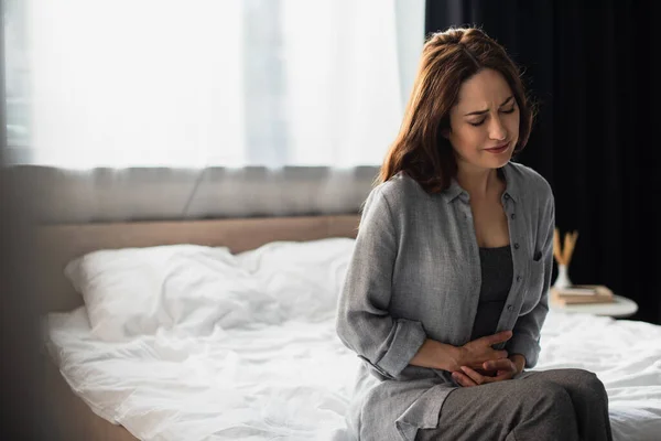 Brunette woman with closed eyes suffering from stomach ache while sitting on bed — Stock Photo