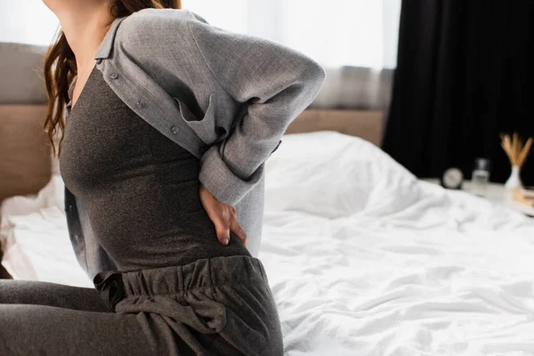 Abgeschnittene Ansicht einer Frau, die auf dem Bett sitzt und unter Rückenschmerzen im Schlafzimmer leidet — Stockfoto