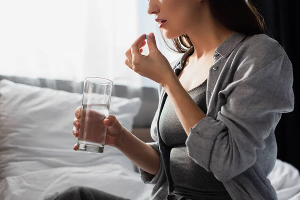 Vista parcial de mujer morena sosteniendo píldora y vaso de agua en casa - foto de stock