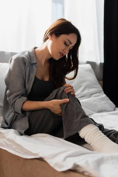 Brunette woman suffering from pain in knee while sitting on bed — Stock Photo