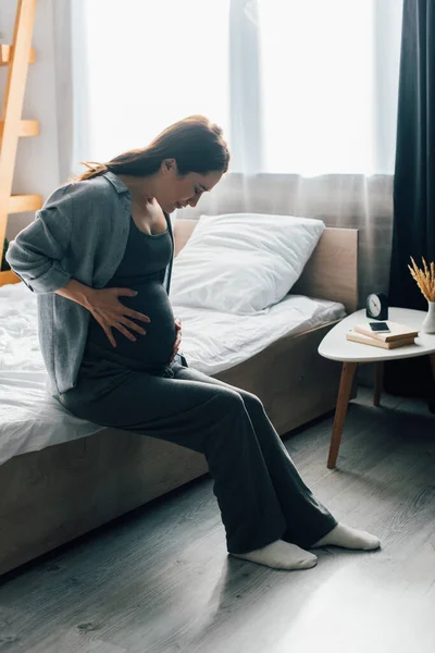 Brunette pregnant woman suffering from pain while sitting on bed — Stock Photo