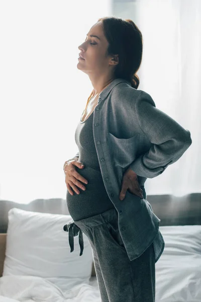 Schwangere leidet unter Schmerzen, während sie Bauch berührt und mit der Hand auf Hüfte im Schlafzimmer steht — Stockfoto