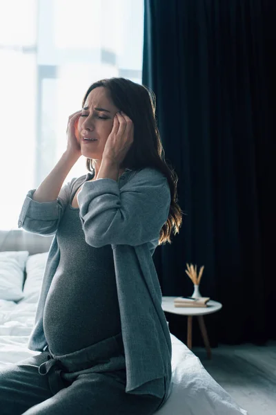Upset pregnant woman touching head while sitting in bedroom — Stock Photo