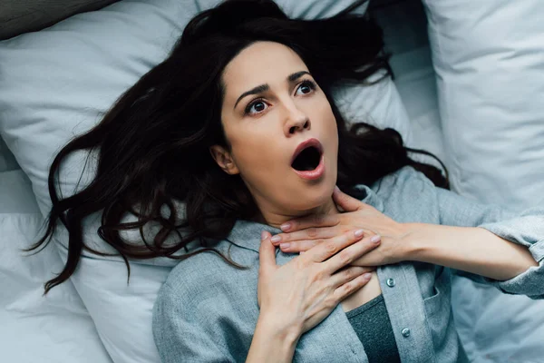Top view of scared brunette woman touching throat while lying on bed — Stock Photo