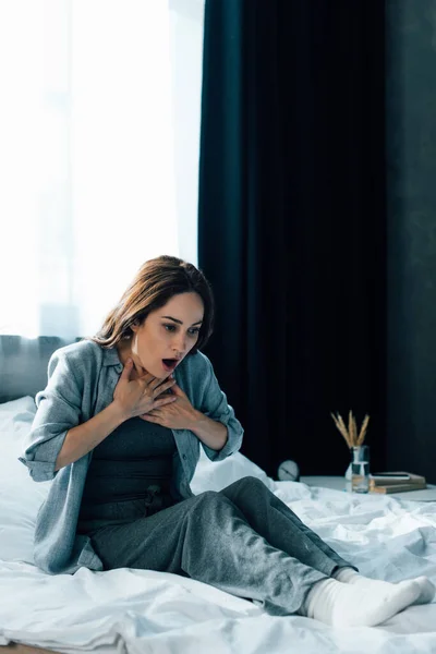 Verängstigte brünette Frau berührt Kehle, während sie auf dem Bett sitzt — Stockfoto