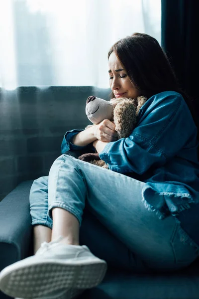 Upset brunette woman with closed eyes holding teddy bear while sitting on sofa — Stock Photo