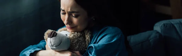 Panoramic crop of sad brunette woman with closed eyes holding teddy bear in living room — Stock Photo