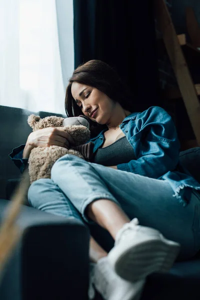 Enfoque selectivo de mujer morena molesta con los ojos cerrados sosteniendo oso de peluche mientras está sentado en el sofá - foto de stock