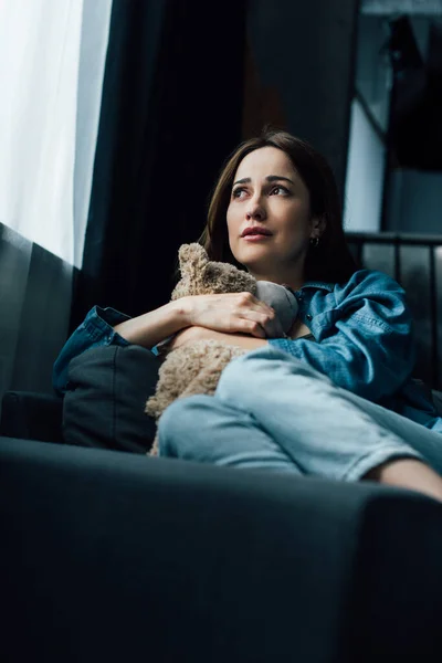 Selective focus of depressed brunette woman holding teddy bear while sitting on sofa — Stock Photo
